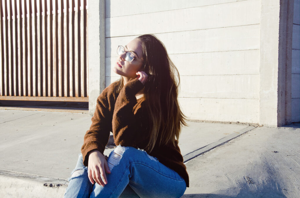 Laura Adriana photo a thoughtful teenager in a brown sweater and blue jeans sitting on a concrete step against a white wall with vertical wooden slats.